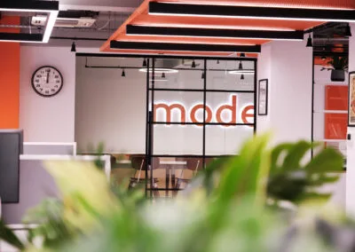 A view through green plants across the office floor to the illuminated branded signage for mode transport in their newly finished Birmingham office - captured by Birmingham property photographer for Copper & Ash Designs and SKK Interiors